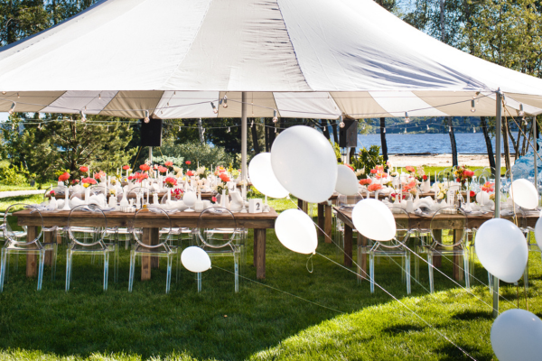 A white party tent setup outdoors covering banquet tables, surrounded by white balloons. 